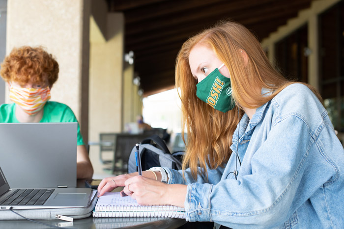 Two masked students seated outdoors with laptops.