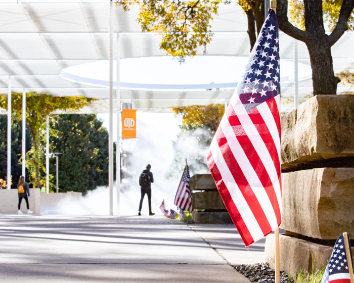 Small American flag along campus sidewalk; McDermott mall and fountain. 